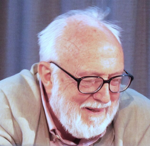 portrait of older man with white beard and hair and glasses looking down and talking